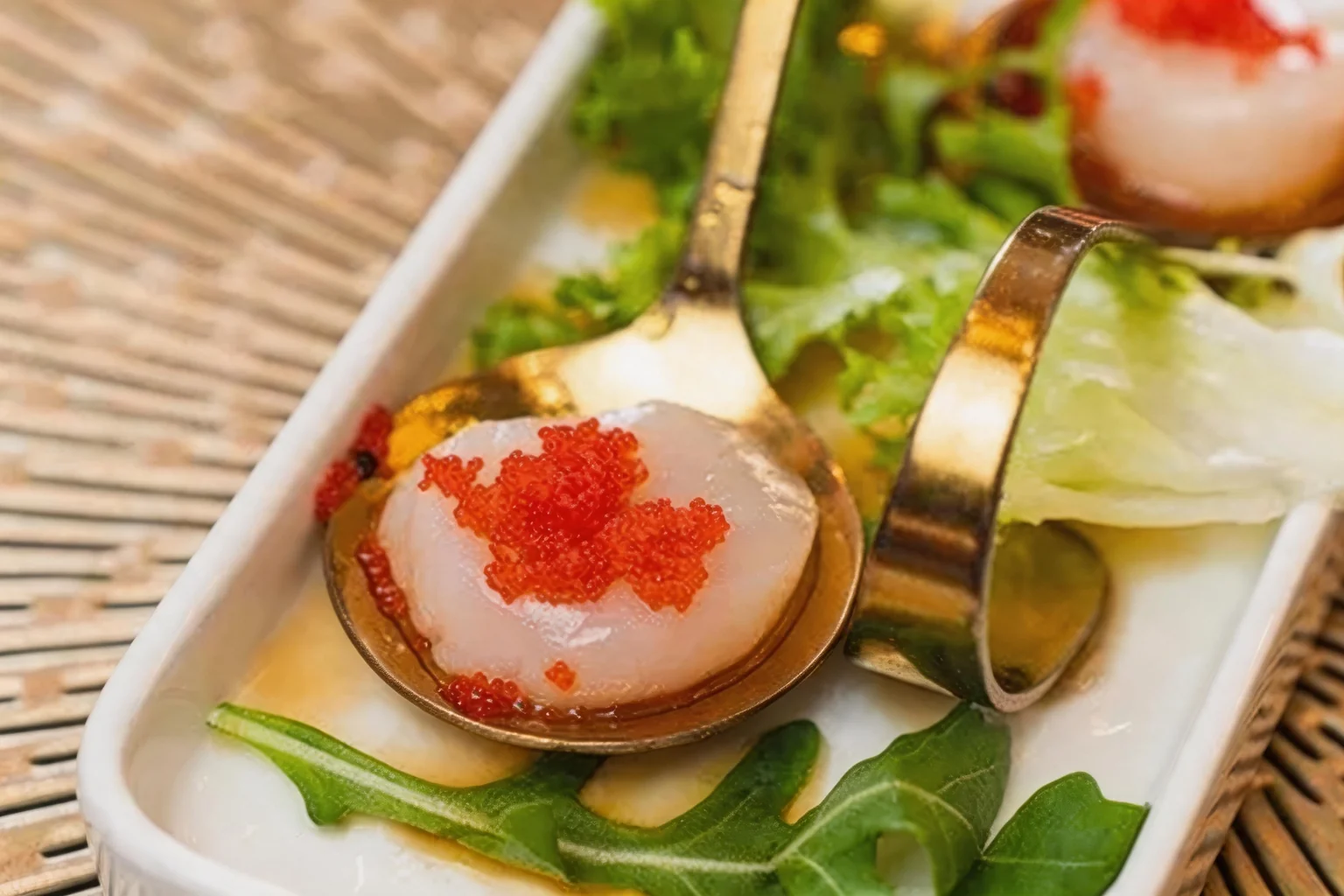 Capesante con tobiko servite in cucchiai dorati, su un piatto bianco con insalata.